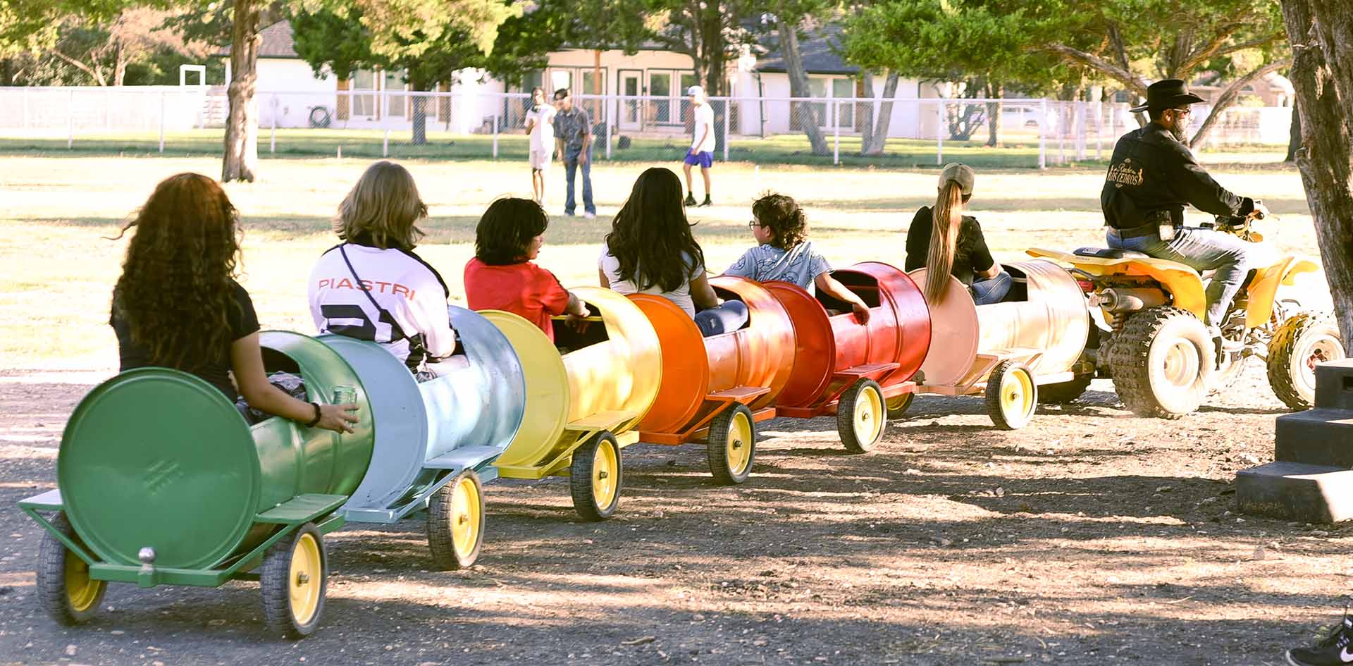 Children and adults riding in colorful barrel train cars pulled by an ATV driven by a cowboy at a ranch petting zoo, highlighting a fun and family-friendly activity for event guests at Rancho Los Cedros